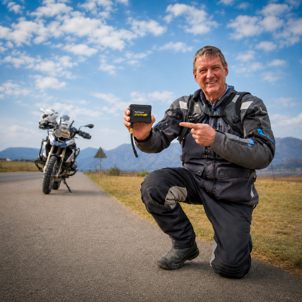 Motorcycle rider holding and pointing to the MotoPressor SmartPUMP cordless tyre inflator beside his adventure motorcycle